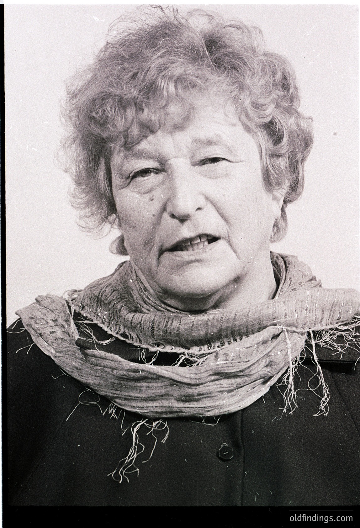 Portrait of an elderly woman in mid-speech, wearing a textured scarf and dark jacket. Black-and-white, high-contrast style suggests vintage photography, likely 1960s–1980s. Expression conveys intensity, possibly for documentary or historical context.