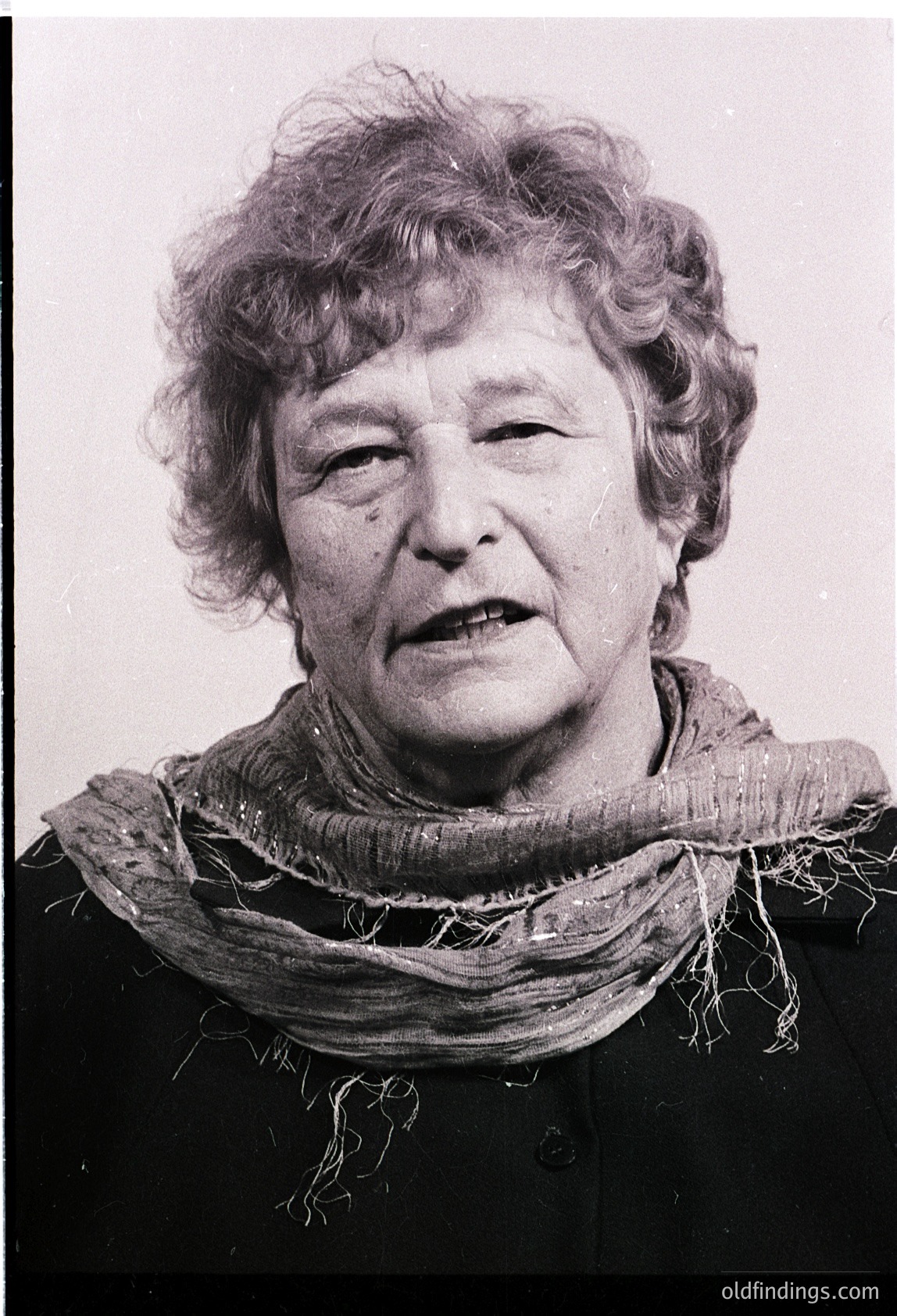 Portrait of an elderly woman in a high-contrast black-and-white photo. She wears a frayed, textured scarf and a dark top, with a neutral expression. Likely mid-20th century (1950s–1970s) based on styling and grain.