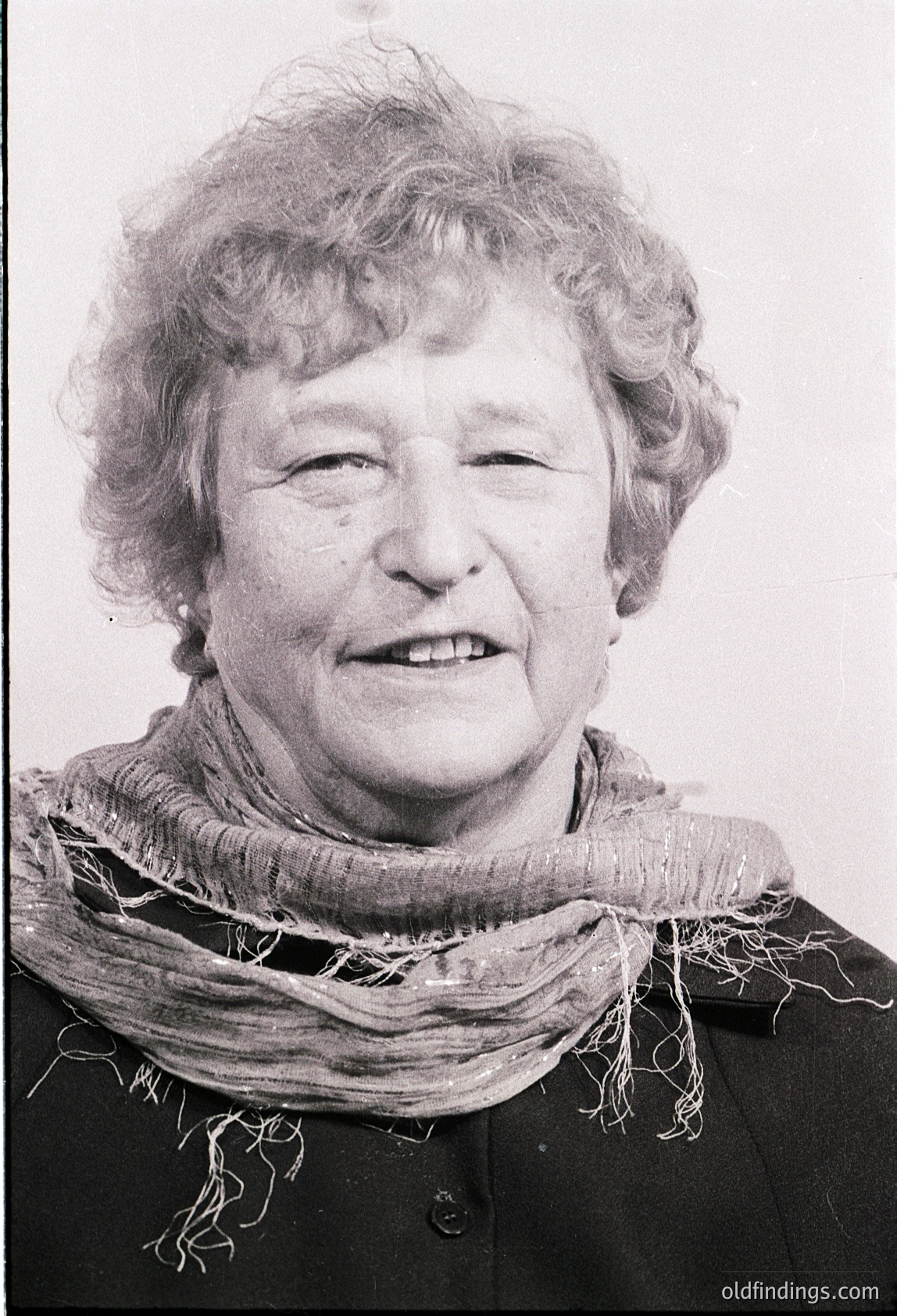 Portrait of an elderly woman with short, wavy gray hair, smiling directly at the camera. She wears a dark jacket with a textured beige scarf draped around her neck. Style suggests mid-20th century (1950s–1970s).