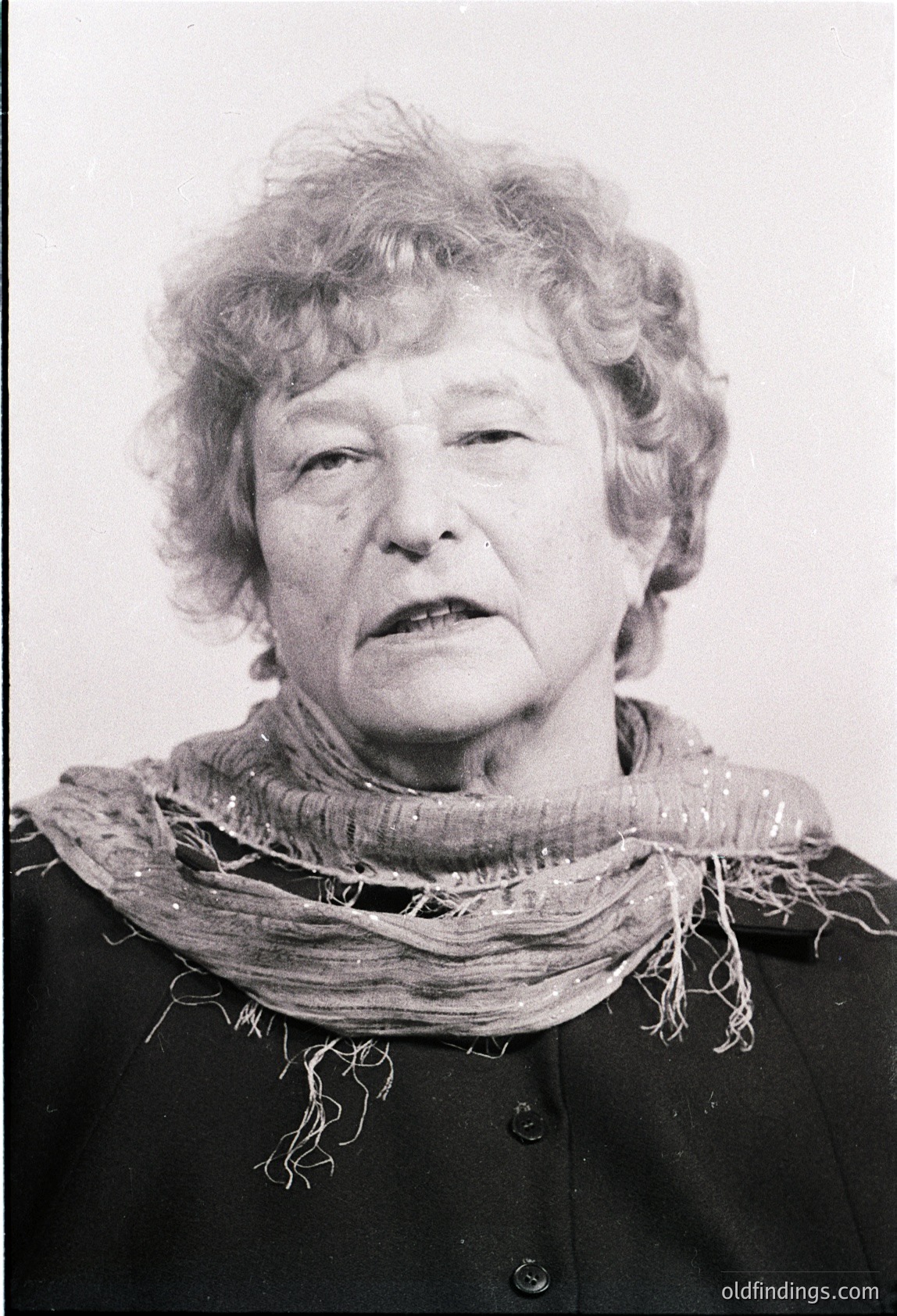 Black-and-white portrait of an elderly woman with short, curly white hair, wearing a textured scarf and dark coat. Expression suggests resilience. Likely mid-20th century (1950s–1970s) based on styling and monochrome.