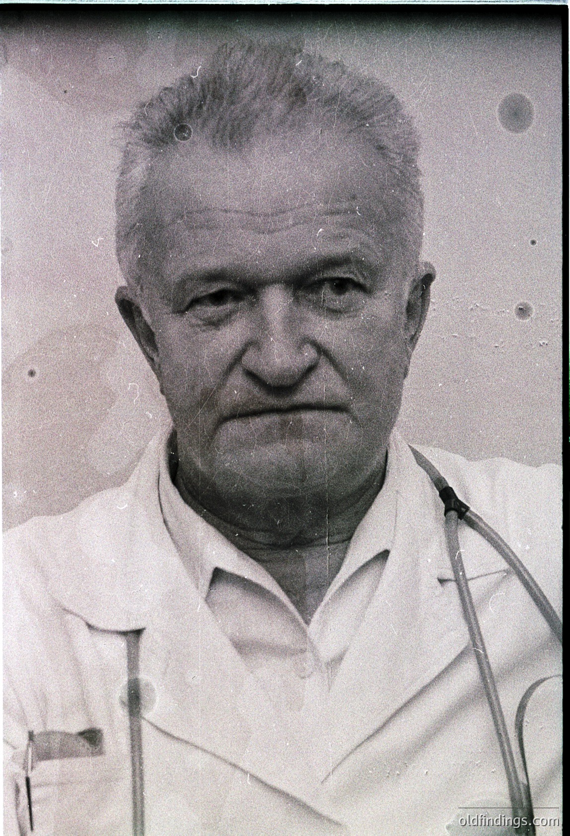 Mid-century black-and-white portrait of an older man in a white lab coat, holding a stethoscope. The grainy, aged film suggests a 1950s–1970s medical setting. Likely a physician or researcher.