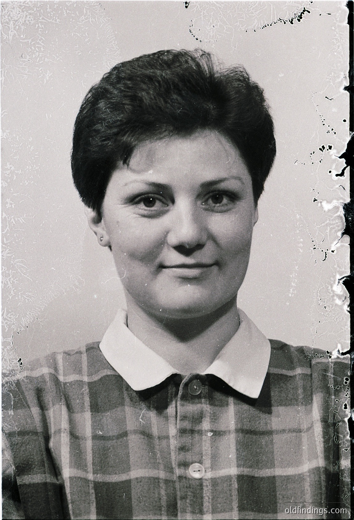 Portrait of a woman in a mid-century plaid blouse, likely from the 1960s–1970s. Short, neatly styled hair and minimalist earrings suggest a formal or identification-style photo. Vintage sepia tone and slight edge wear indicate archival or historical use.