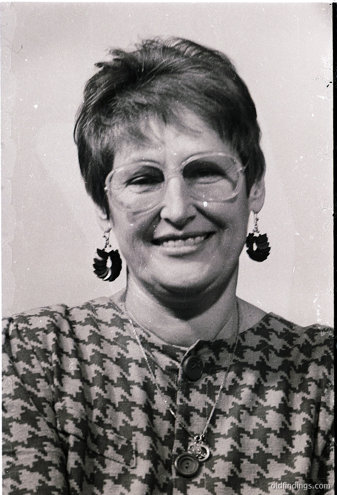 Portrait of a woman in a patterned blouse with large, geometric earrings and a decorative brooch. Mid-20th century fashion, likely 1960s–1970s. Soft focus and warm lighting enhance vintage charm.