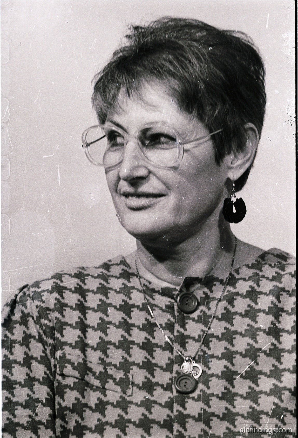 Portrait of a woman in a patterned blouse with geometric checkered design, wearing round-framed glasses and large hoop earrings. Necklace features a circular pendant. Likely mid-20th century (1960s–1970s) studio portrait.