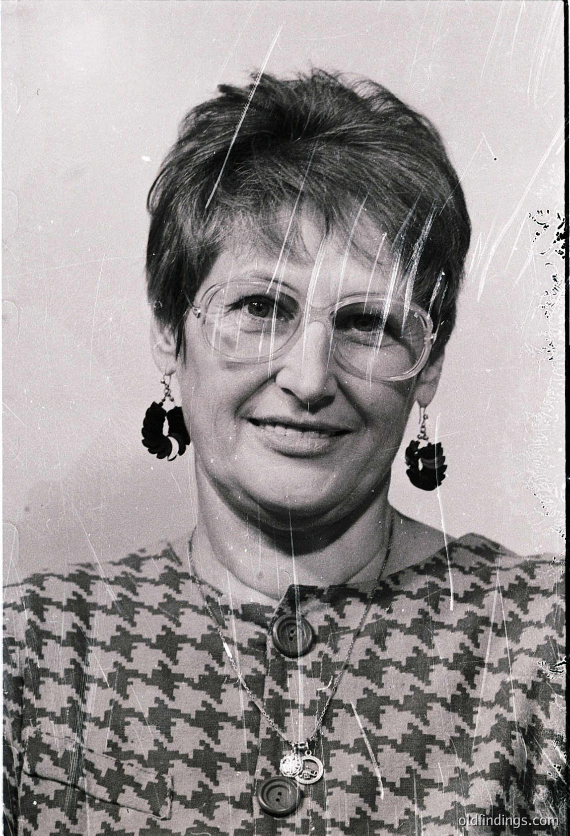 Portrait of a woman in mid-20th century fashion, wearing round-framed glasses and large, geometric-pattern earrings. Her patterned blouse features a prominent pin or badge. Likely or European style.