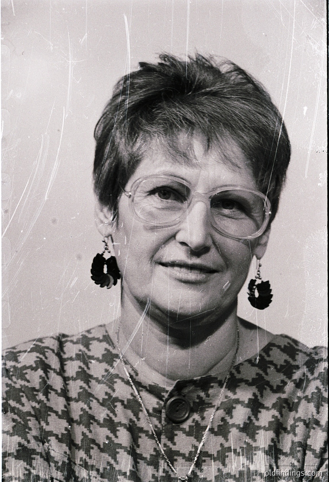Portrait of a woman in a patterned blouse, short hairstyle, and round glasses, wearing distinctive black earrings with circular motifs. Black-and-white photo with slight grain, suggesting mid-20th century (1950s–1970s). Neutral background suggests studio or formal setting.