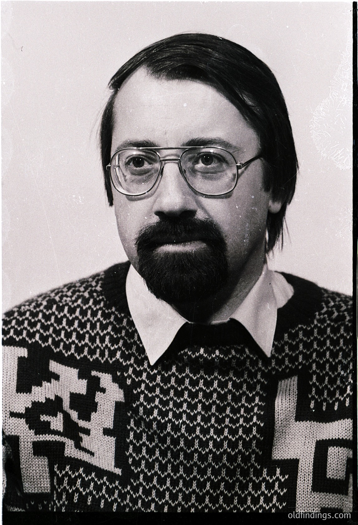 Portrait of a man in a patterned sweater, likely from the 1970s–1980s. Geometric, geometric-textured knit with bold black-and-white contrast. Glasses with thin metal frames. Neutral expression, plain background.