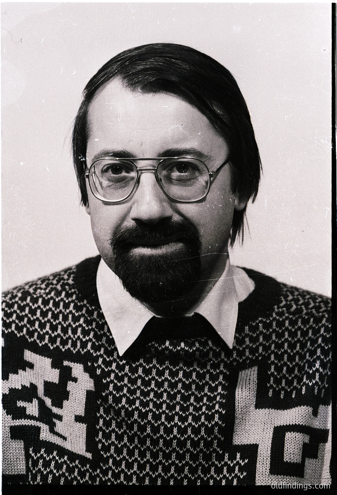 Portrait of a man in a patterned sweater with geometric motifs, likely 1970s-1980s. Glasses and short beard frame a neutral expression. Plain background suggests studio or formal setting.
