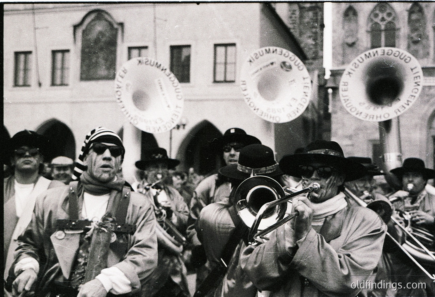 Vintage black-and-white street band in traditional European attire, playing brass instruments and cymbals. Uniforms feature peaked caps, scarves, and ornate buttons. Architectural backdrop includes a building with arched windows and a banner reading *"Temeschwarer Musikverein."* Likely late 19th to early 20th century, Central/Eastern Europe.