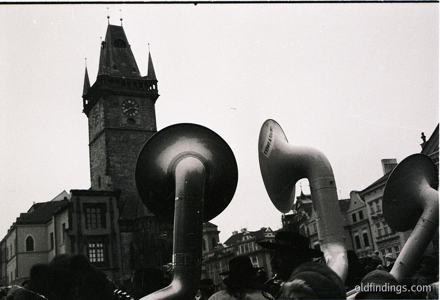 Black-and-white street scene featuring large megaphones in foreground, likely from a public gathering or rally. Prominent clock tower with Gothic Revival architecture in background, suggesting European setting. Crowd partially obscured by megaphones, indicating organized event. Style and architecture hint at mid-20th century (1940s–1960s).