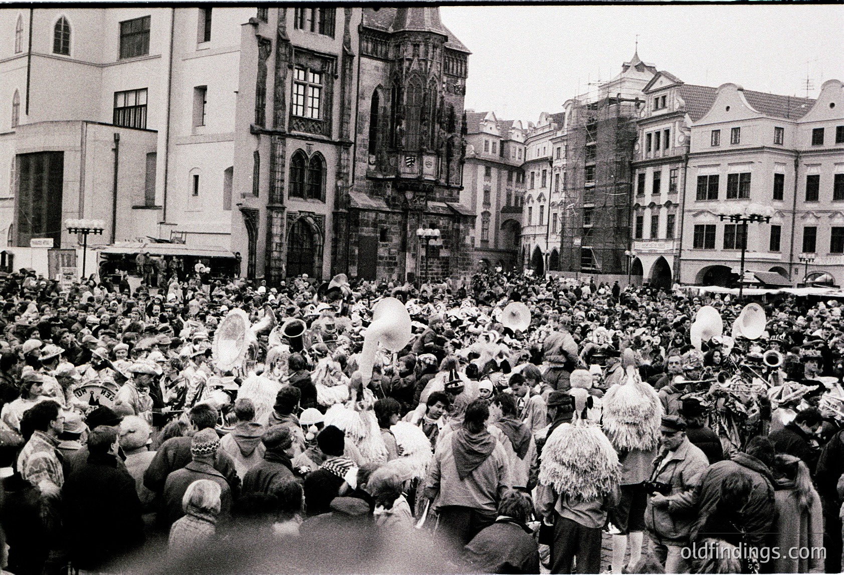 Crowded European town square with Baroque-style architecture, featuring ornate facades and a clock tower. Mid-20th century (likely 1950s–60s) gathering of people in winter attire—fur hats, coats, and scarves—suggesting a festive or public event. Dense urban setting with historic buildings and a lively atmosphere.