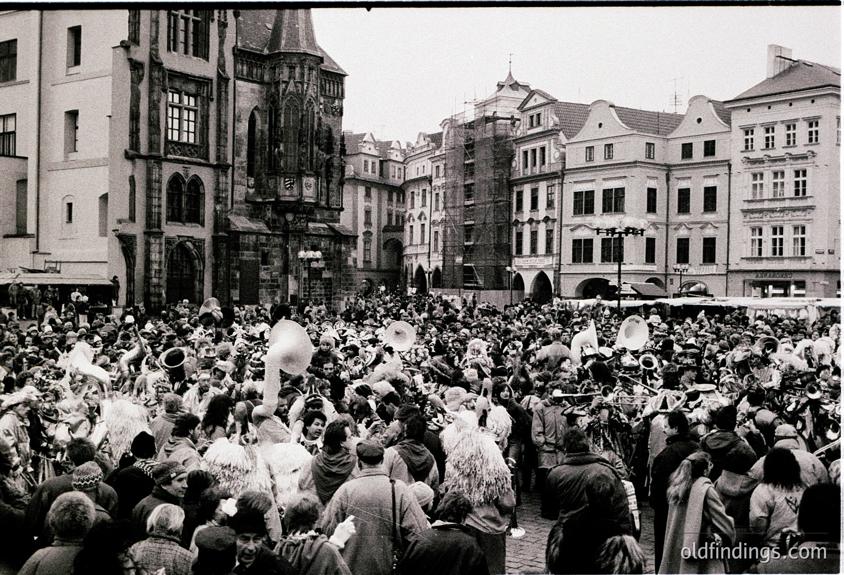 Historic street festival in Prague’s Old Town Square, featuring Baroque/Renaissance architecture with ornate facades and arched gateways. Dense crowd in 1960s-era attire, including fur hats and coats, likely during winter carnival or holiday celebration. Crowd density and attire suggest mid-20th century European cultural event.