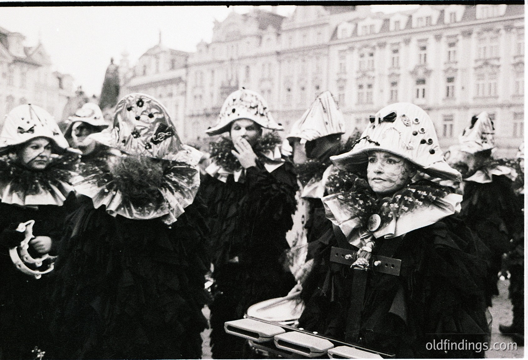Black-and-white street scene featuring women in elaborate, ornate carnival masks and fur-trimmed coats, likely from a 19th–early 20th century European festival. Architectural backdrop suggests a grand urban plaza with classical buildings. Mask designs include exaggerated features, feathers, and decorative embellishments.