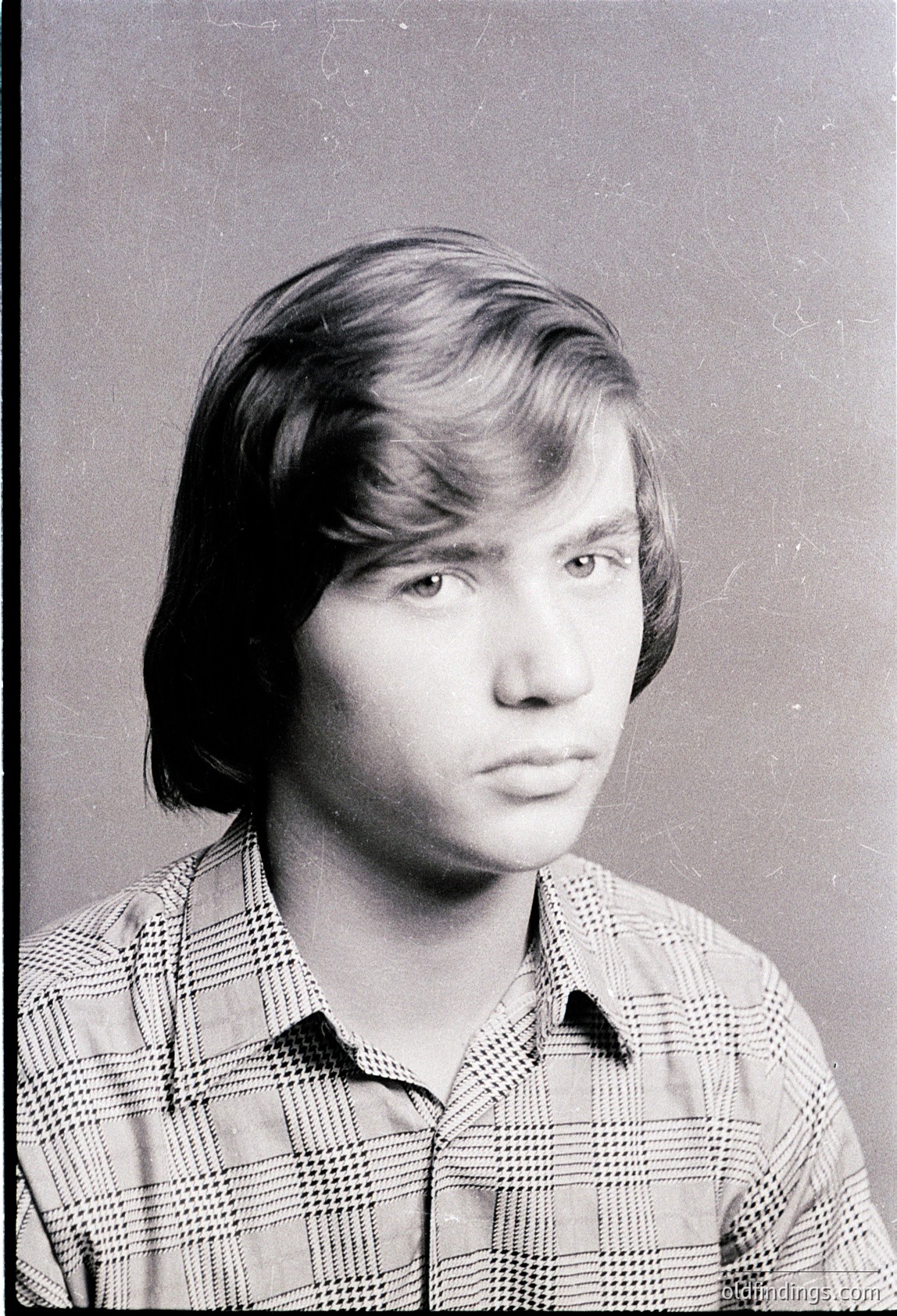 Portrait of a young man in a plaid button-up shirt, likely a 1970s school or ID photo. Neutral expression, short side-swept hair. Plain studio background suggests institutional use.