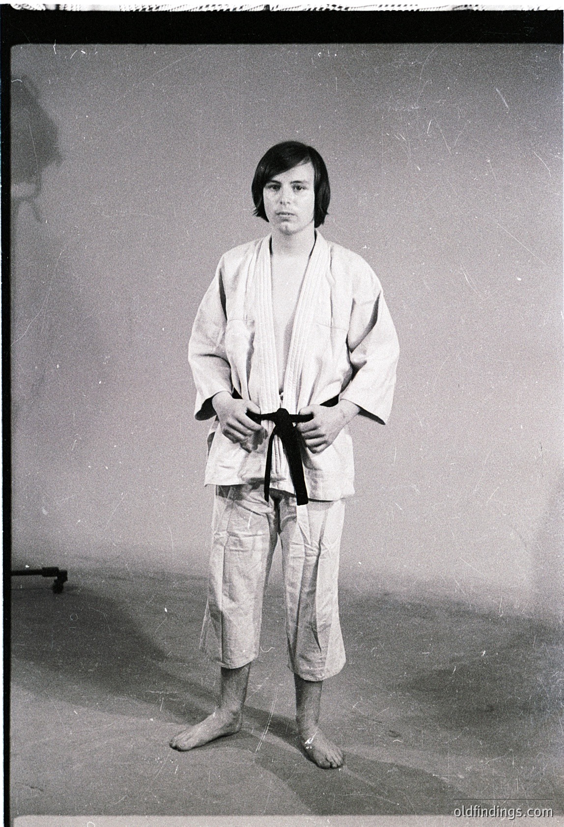 Portrait of an individual in a traditional judogi (karate/judo uniform) with a black belt, likely mid-20th century. Plain indoor studio background suggests formal or instructional setting. Distinctive hairstyle and clothing indicate martial arts training or competition.