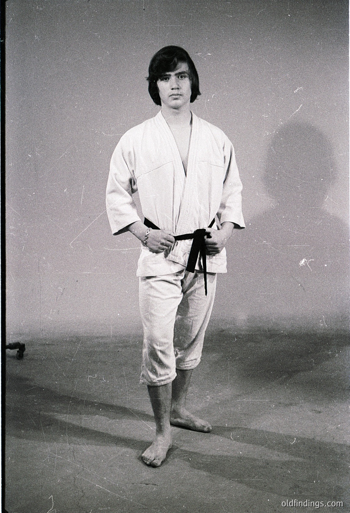 Portrait of a person in traditional judo attire (gi), standing indoors with neutral background. Belt appears to be black or dark brown, suggesting rank. Mid-20th century styling (1950s–1970s) with short haircut. Ideal for martial arts history, vintage sports, or cultural studies research.