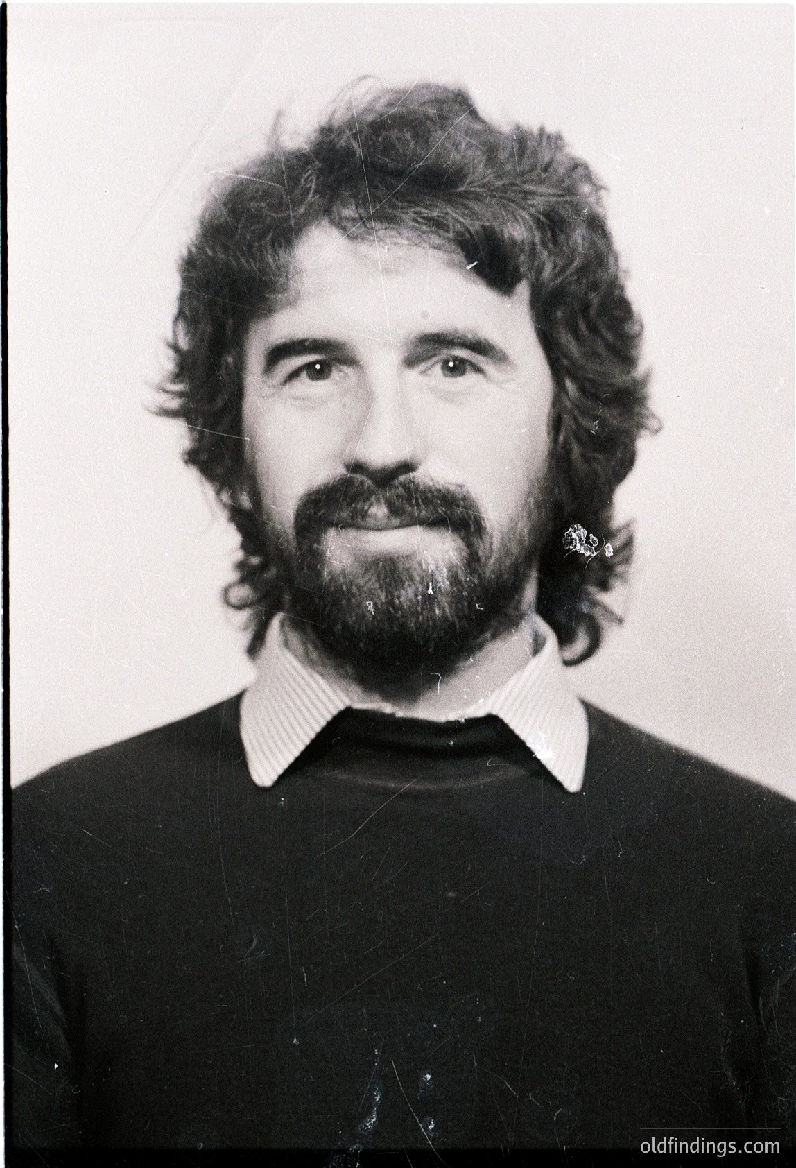 Portrait of a man with long sideburns, mustache, and beard, wearing a dark sweater and striped collared shirt. Classic 1970s-1980s male grooming style. Plain background suggests studio or ID photo.