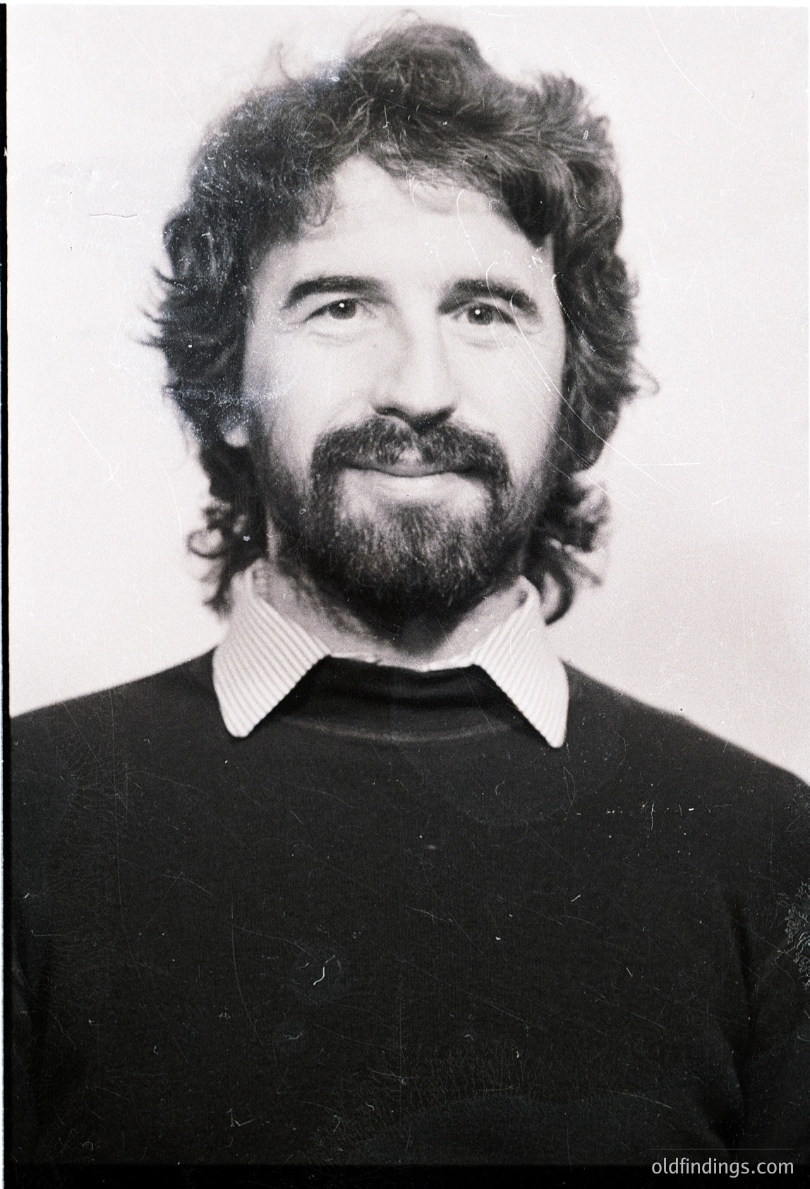 Portrait of a man with a full beard and mustache, wearing a striped collared shirt under a dark sweater. Mid-length, wavy hair styled back. Black-and-white studio photo, likely 1970s–1980s.