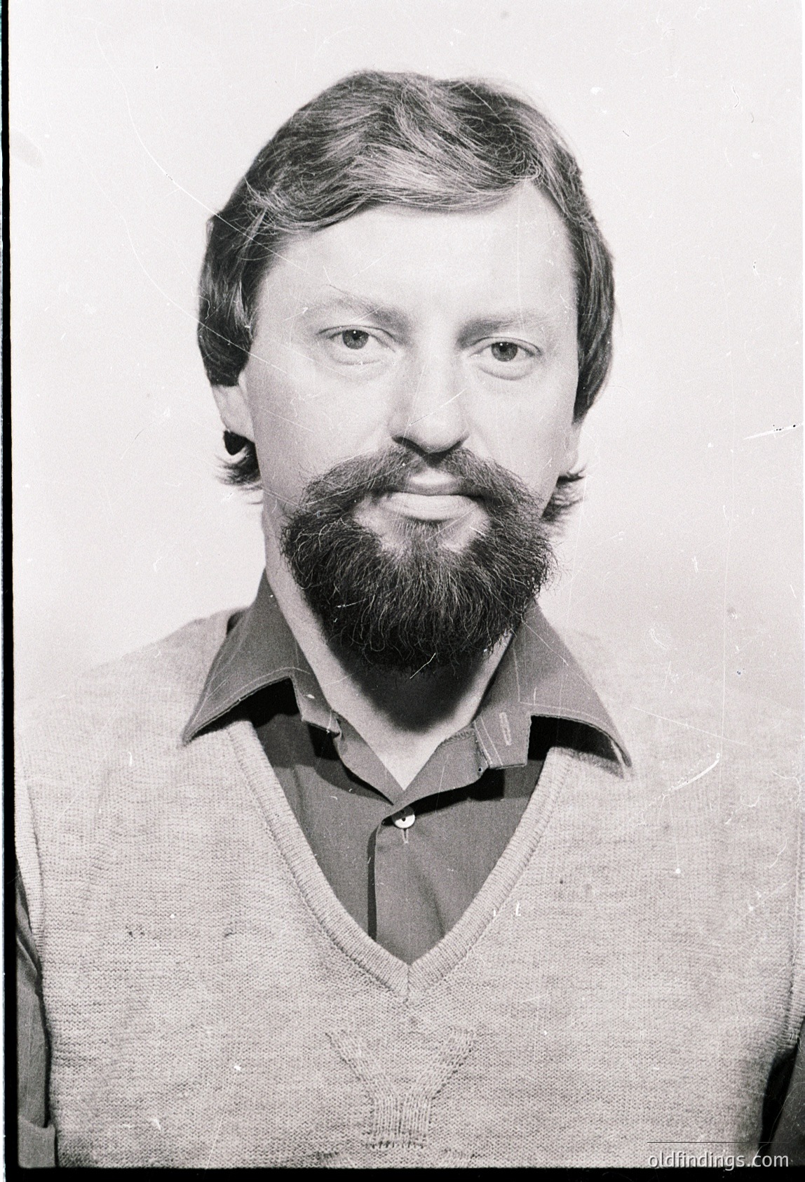 Portrait of a man with a full beard and mustache, wearing a V-neck sweater. Style suggests 1970s–1980s Western Europe/USA. Neutral expression, likely a professional or identification photo.