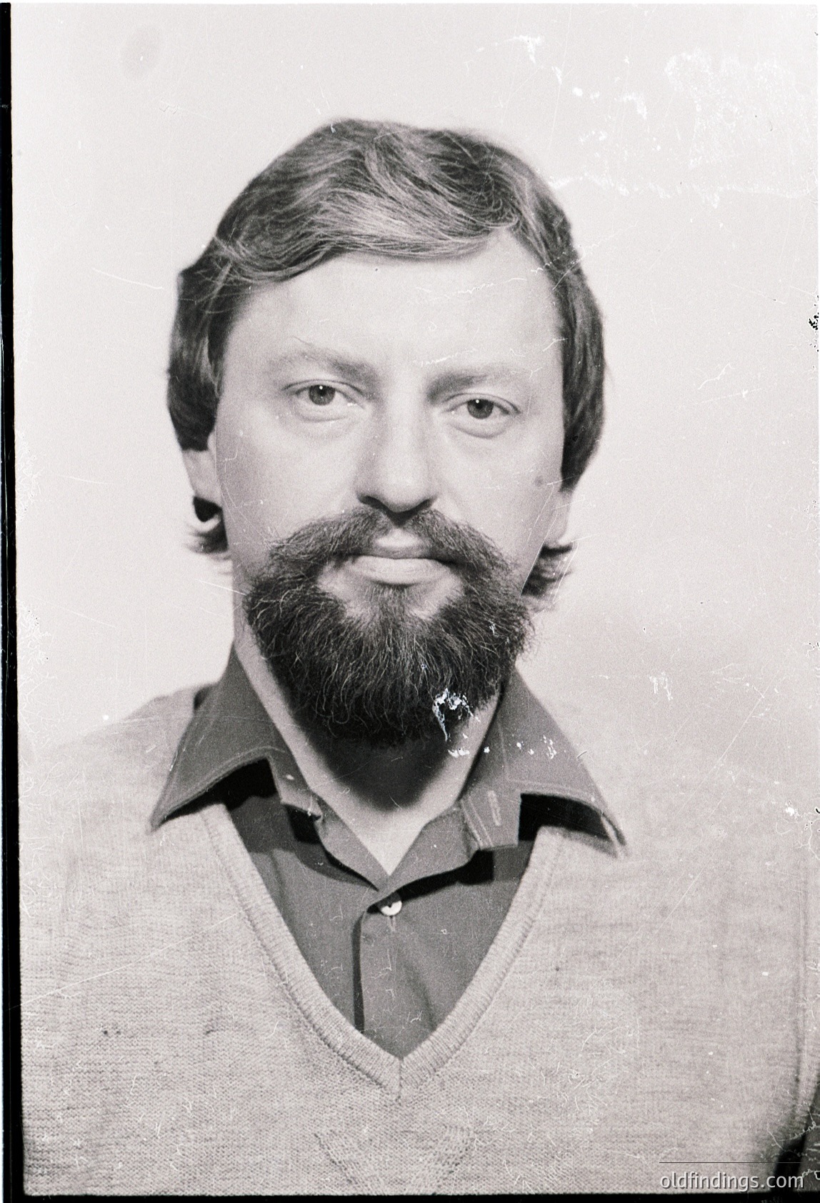 Portrait of a man with a full beard and mustache, wearing a collared V-neck sweater. The style suggests a mid-20th century look, likely 1960s–1970s. Neutral, indoor lighting with a plain background.