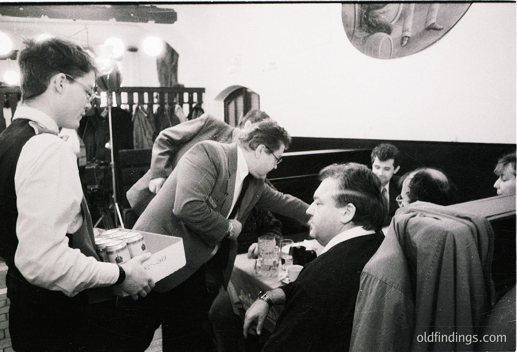 Vintage black-and-white photo of a group of men in formal attire (suits, ties) playing chess in a dimly lit room. One man in a white shirt and glasses holds a tray with chess pieces. Others focus intently on their boards. Hanging light fixture and wooden furniture suggest a mid-century institutional or club setting. Likely or ,