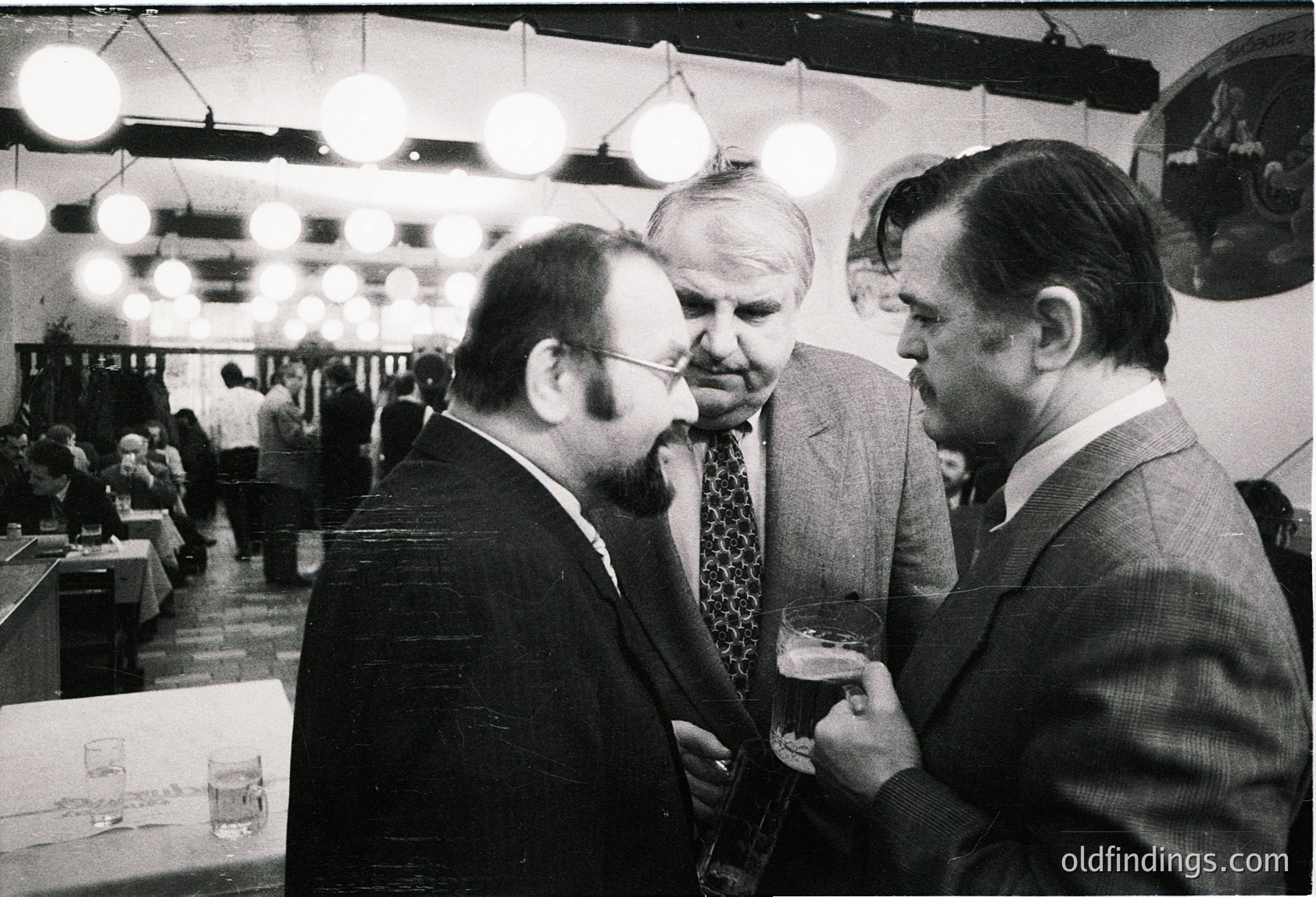 Three men in formal attire—dark suits, ties, and dress shirts—engage in conversation at a mid-20th-century indoor event. Suspended globe lights illuminate the space, while tables and attendees suggest a conference or exhibition hall. The setting reflects midcentury professional culture.