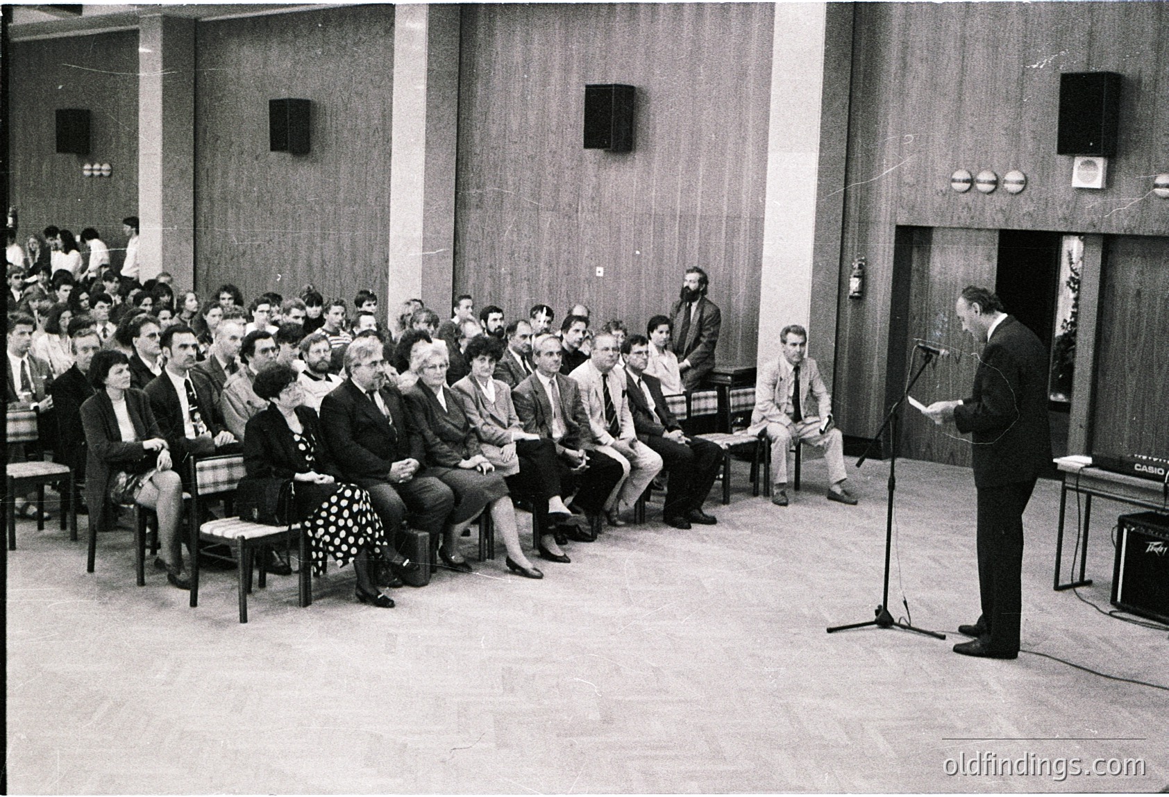 Mid-century lecture hall with tiered seating, wooden paneling, and industrial lighting. A speaker addresses an attentive audience of men in formal attire, likely 1960s–1970s. Microphone stand and chalkboard hint at educational or professional setting.