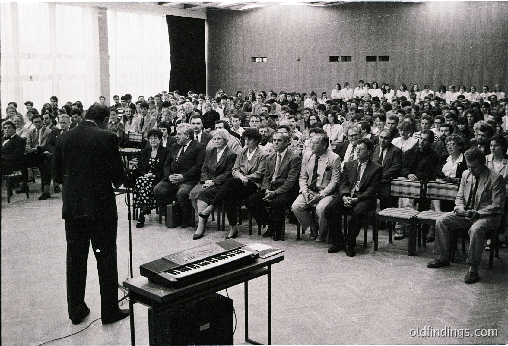 A formal lecture or conference in a mid-century auditorium, featuring a speaker addressing a packed audience seated in tiered rows. The speaker stands near a vintage keyboard amplifier, suggesting a technical or educational presentation. Attendees wear business attire, indicating a professional or academic setting. The setting’s minimalist decor and lighting reflect 1960s–1970s institutional design. [Mid-century auditorium lecture with keyboard amplifier ]