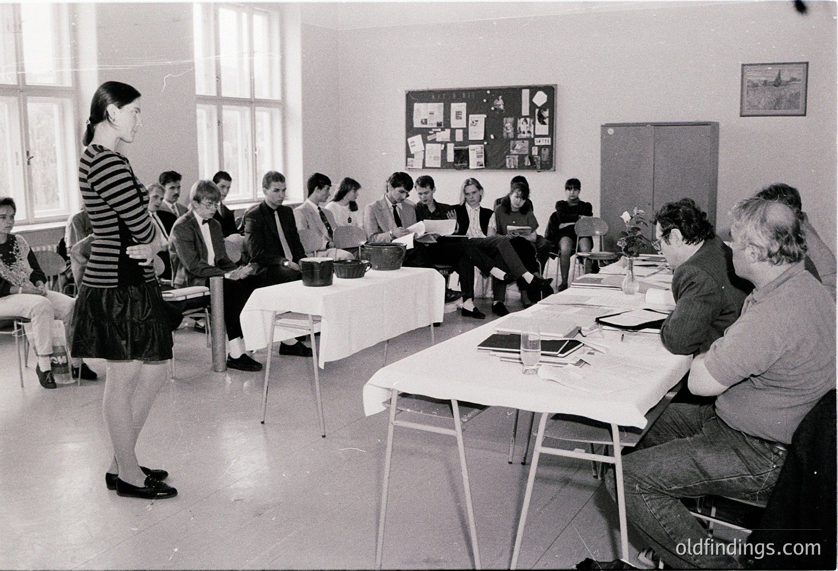 Black-and-white classroom scene featuring a teacher addressing students seated at long tables, 1960s–1970s. Functional, institutional lighting and bulletin board with pinned images. Uniform attire suggests formal education setting.