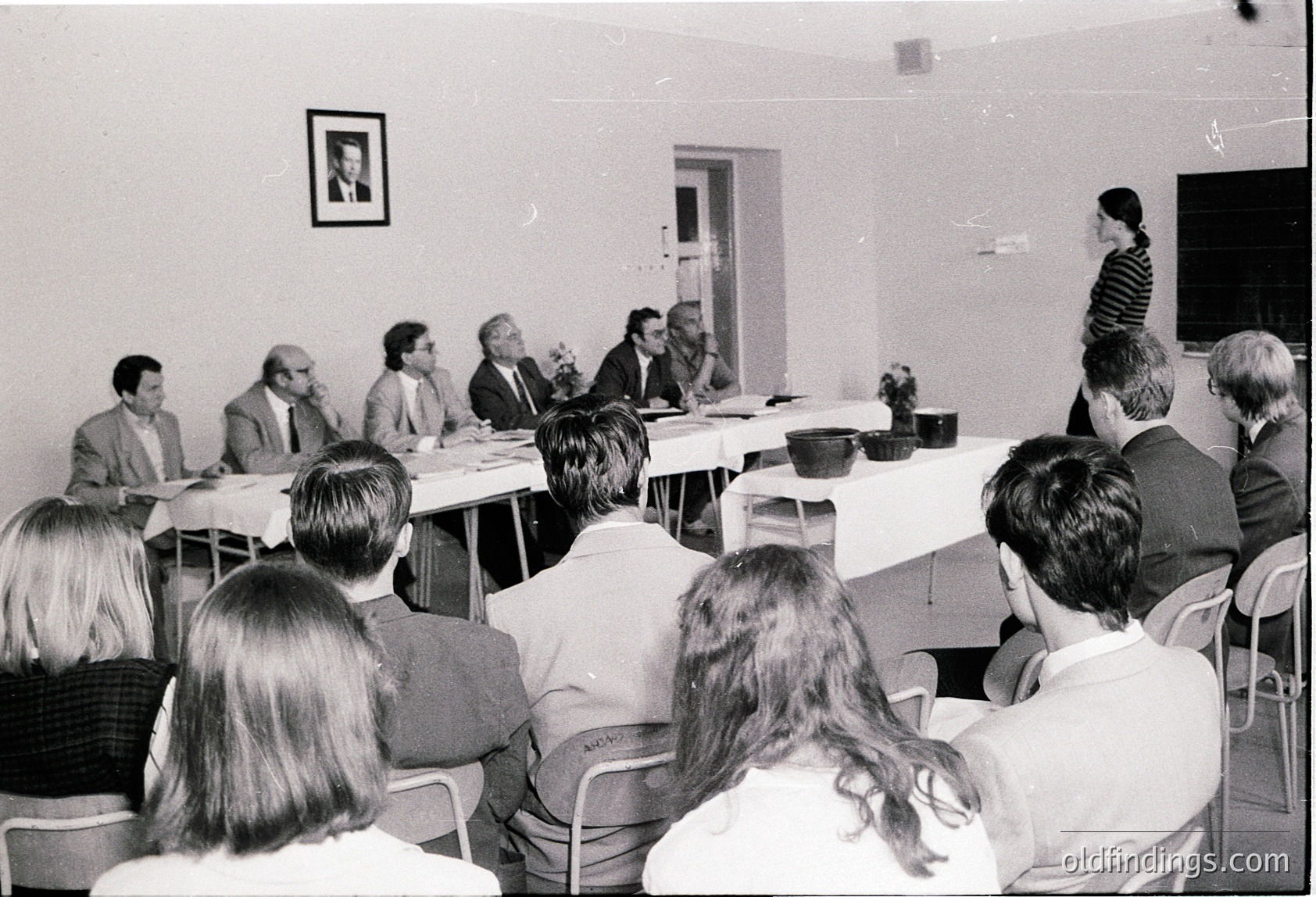 Black-and-white conference scene: seated panelists at a rectangular table in a plain room, with one speaker addressing an audience of seated attendees. Formal attire suggests a mid-20th-century professional or governmental setting. Decor minimal—framed portrait, potted plant, and basic furniture.