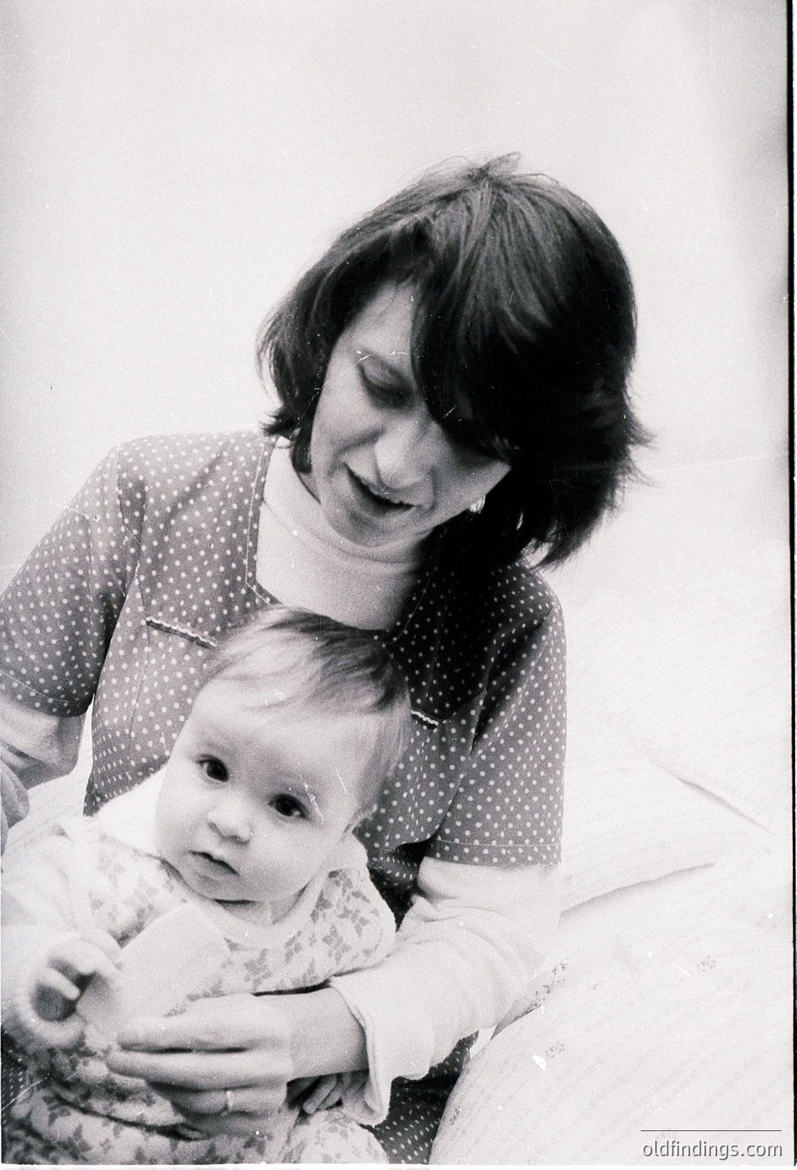 A tender black-and-white portrait of a woman cradling an infant, likely from the 1960s–1970s. The woman wears a polka-dot blouse with a high-necked collar, while the baby is dressed in a floral onesie. Soft indoor lighting enhances the intimate moment. Ideal for vintage family archives or nostalgic design references.