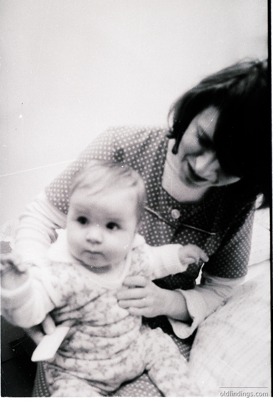 Black-and-white portrait of a woman cradling an infant, likely mid-20th century. Woman wears a polka-dot blouse and a choker necklace; baby holds a stuffed toy. Indoor setting, soft focus. Captures candid warmth of family life.