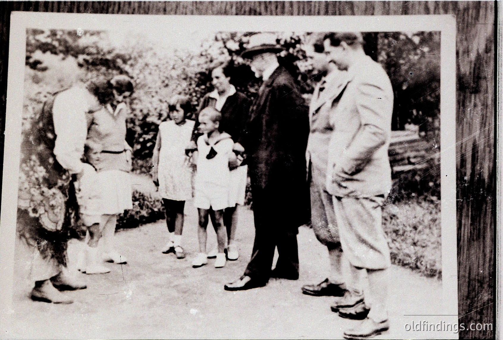 Mid-20th century black-and-white photo of a family group outdoors, likely mid-1950s. Six individuals—adults in long-sleeve shirts and trousers, two children in dresses—pose on a gravel path surrounded by greenery. One adult holds a child, another wears a hat. Formal, candid family portrait style.