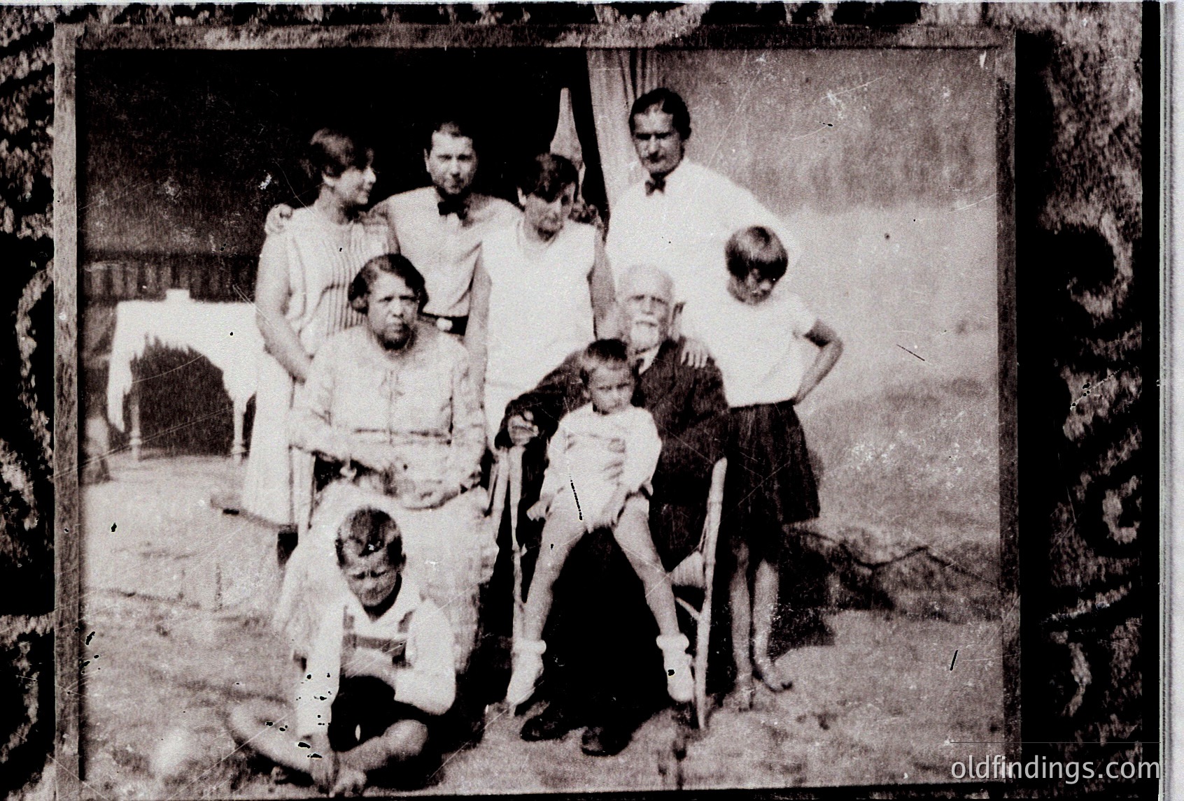 Vintage sepia-toned family portrait, likely late 19th to early 20th century. Eight individuals pose outdoors near a simple wooden structure, with one seated in a wheelchair. Clothing suggests modest, traditional attire.