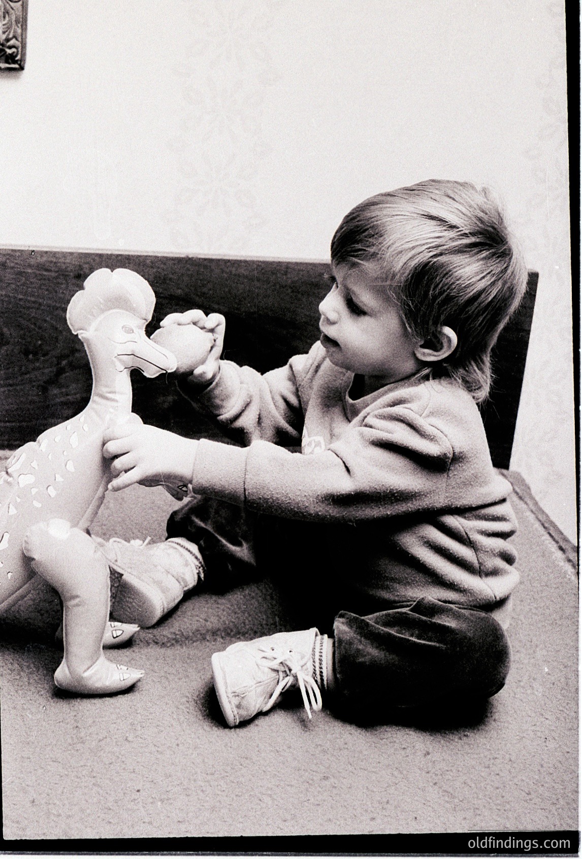 Mid-20th century black-and-white photo of a young child playing with a large ceramic duck toy, seated on a carpeted floor. The child wears a short-sleeved shirt and knee-high socks. Toy features a detailed beak and textured feathers. Likely 1950s–1960s domestic setting.