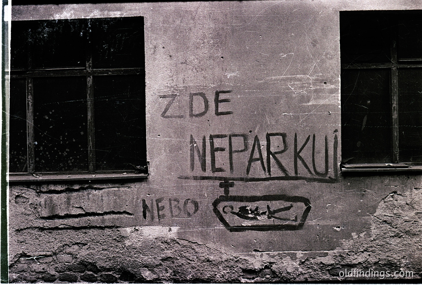 Black-and-white graffiti on weathered concrete wall reads *"ZDE NEPARKUJ!"* ("Don’t park here!") with a crossed-out parking symbol below. Cracked plaster and peeling paint suggest aged urban infrastructure. Likely Eastern European, mid-20th century.