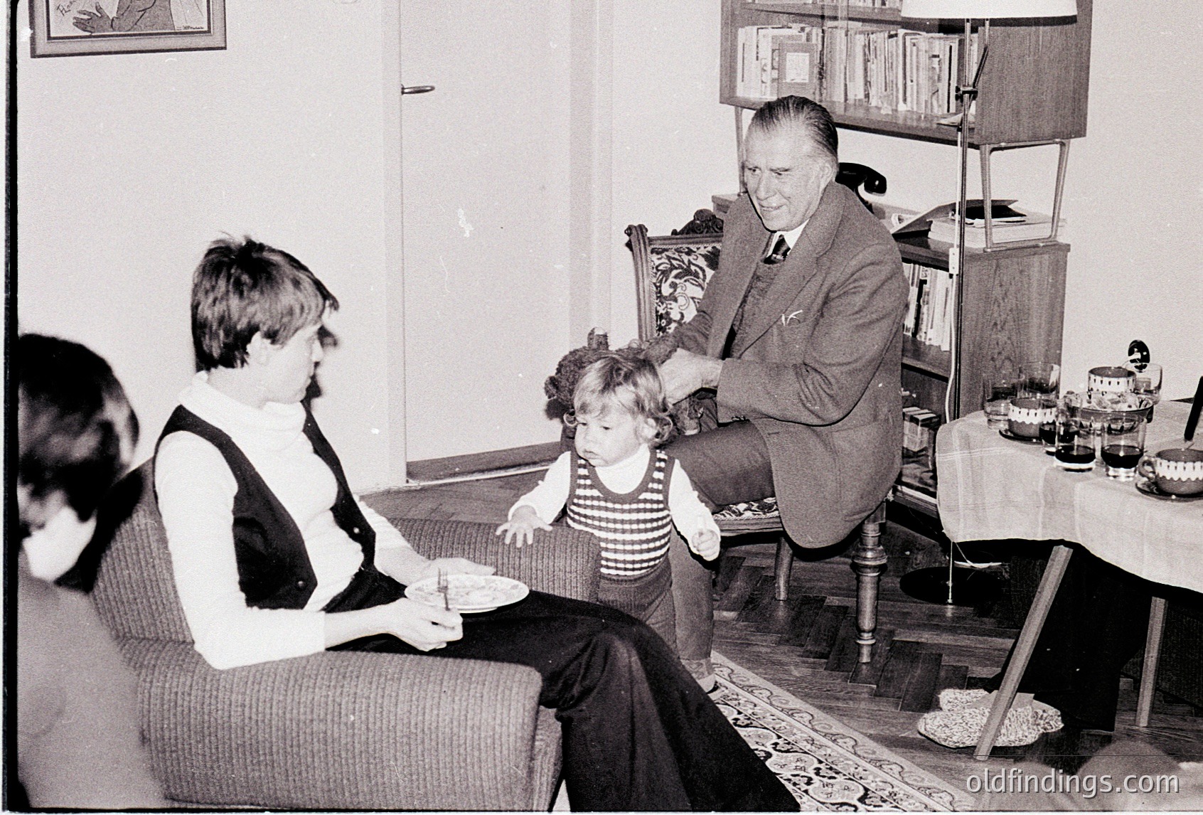 Black-and-white interior shot of a mid-20th-century home, likely 1960s–1970s. A man in formal attire (vest, tie) interacts with a toddler in a striped dress, while a woman in a blouse sits beside them. Wooden furniture, bookshelves, and a round table with decorative plates suggest a middle-class household. Warm, intimate family moment captured.