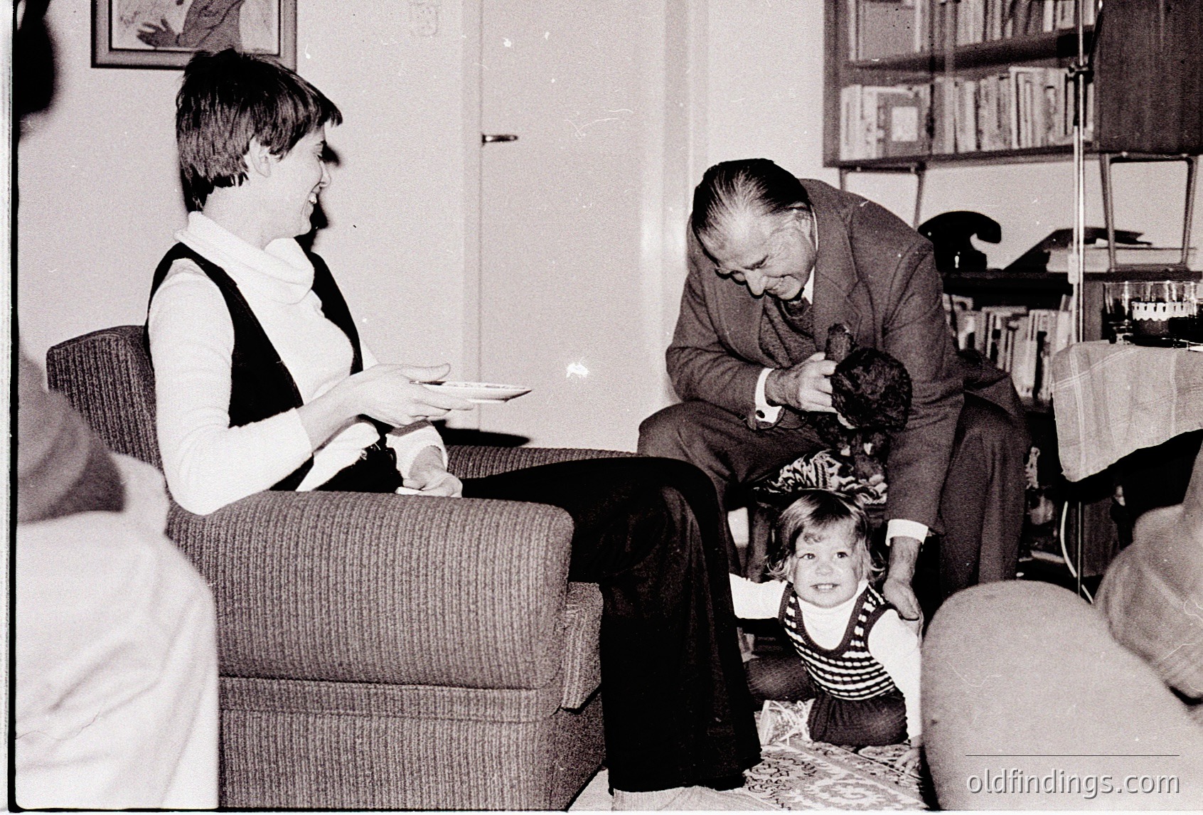 Mid-20th century living room scene: a woman in a turtleneck sweater holds a tray, an elderly man in suit and tie interacts with a toddler in striped shirt. Bookshelves and vintage furniture suggest a midcentury home.