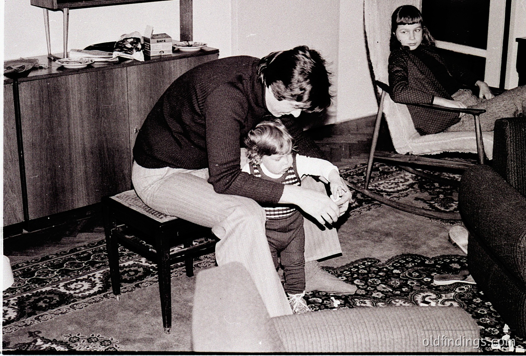 A tender 1970s domestic scene: an adult kneels beside a young child on a wooden stool, offering comfort. A second child sits on a rug near a patterned carpet, while a wooden cabinet and chair frame the background. Warm, intimate family moment.