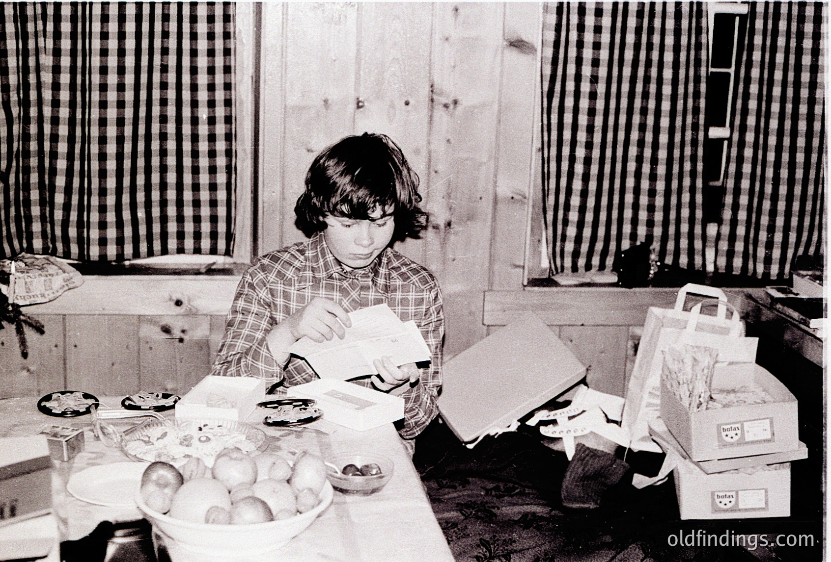 A young person in a plaid shirt organizes paper packages on a cluttered wooden table surrounded by household items. Fresh fruit bowl and cardboard boxes with labels in the background. Likely a mid-20th century domestic scene, possibly or .