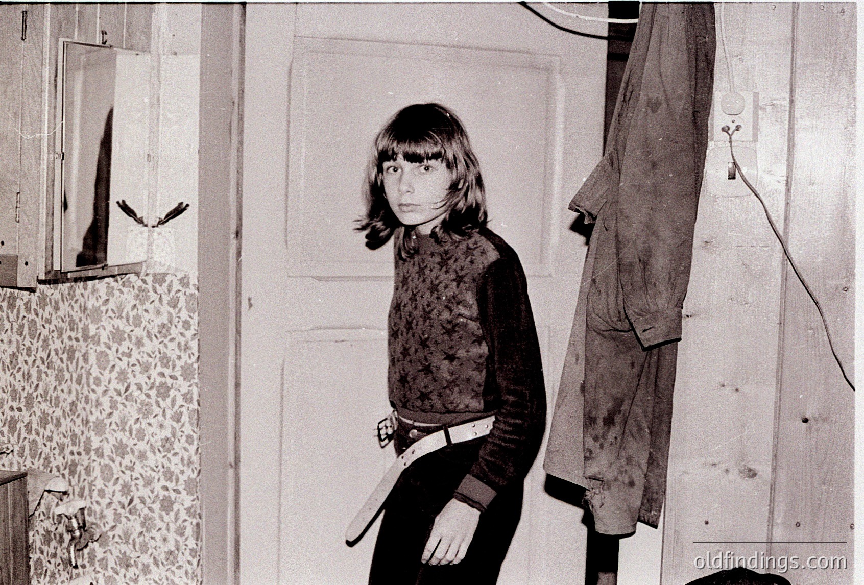 A young girl in a patterned sweater stands in a dimly lit indoor space, holding a belt. The wallpaper features floral patterns, and a coat hangs on a hook beside her. Likely mid-20th century, possibly Eastern Europe.
