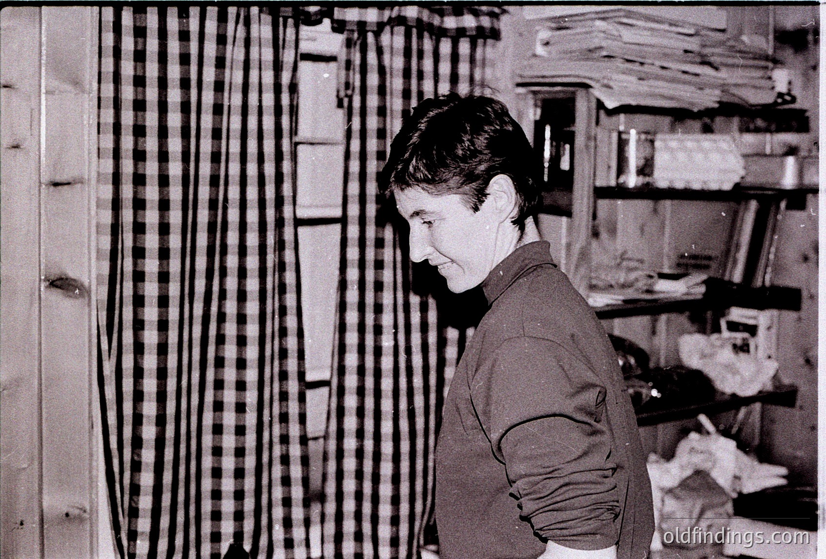 Black-and-white portrait of a person standing in a textile workshop, surrounded by stacked fabric rolls and woven textiles. The geometric checkered pattern in the background suggests handwoven or industrial textile production. Likely Eastern European, 1960s–1980s.