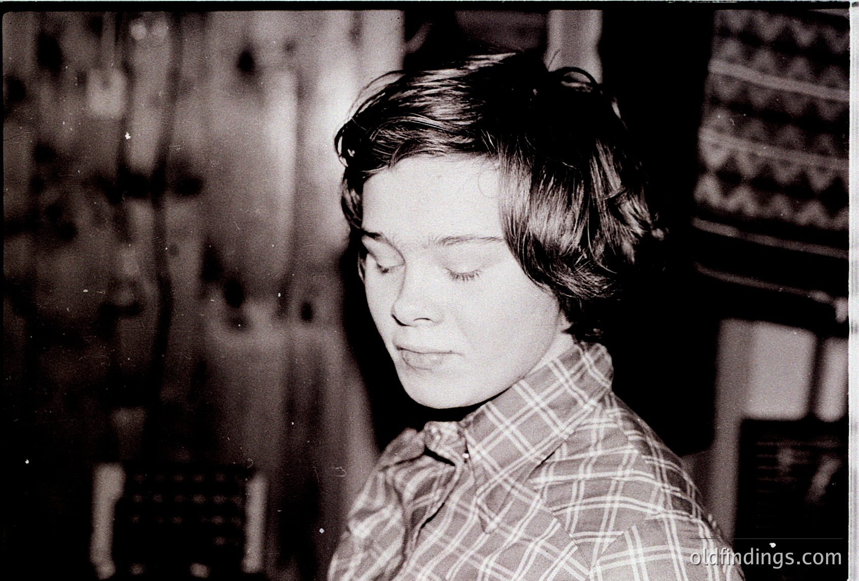 Portrait of a young woman in a plaid shirt, captured in black-and-white. Her short, textured hair and contemplative expression evoke mid-century introspection. Background suggests an indoor setting with patterned wallpaper and blurred furnishings.