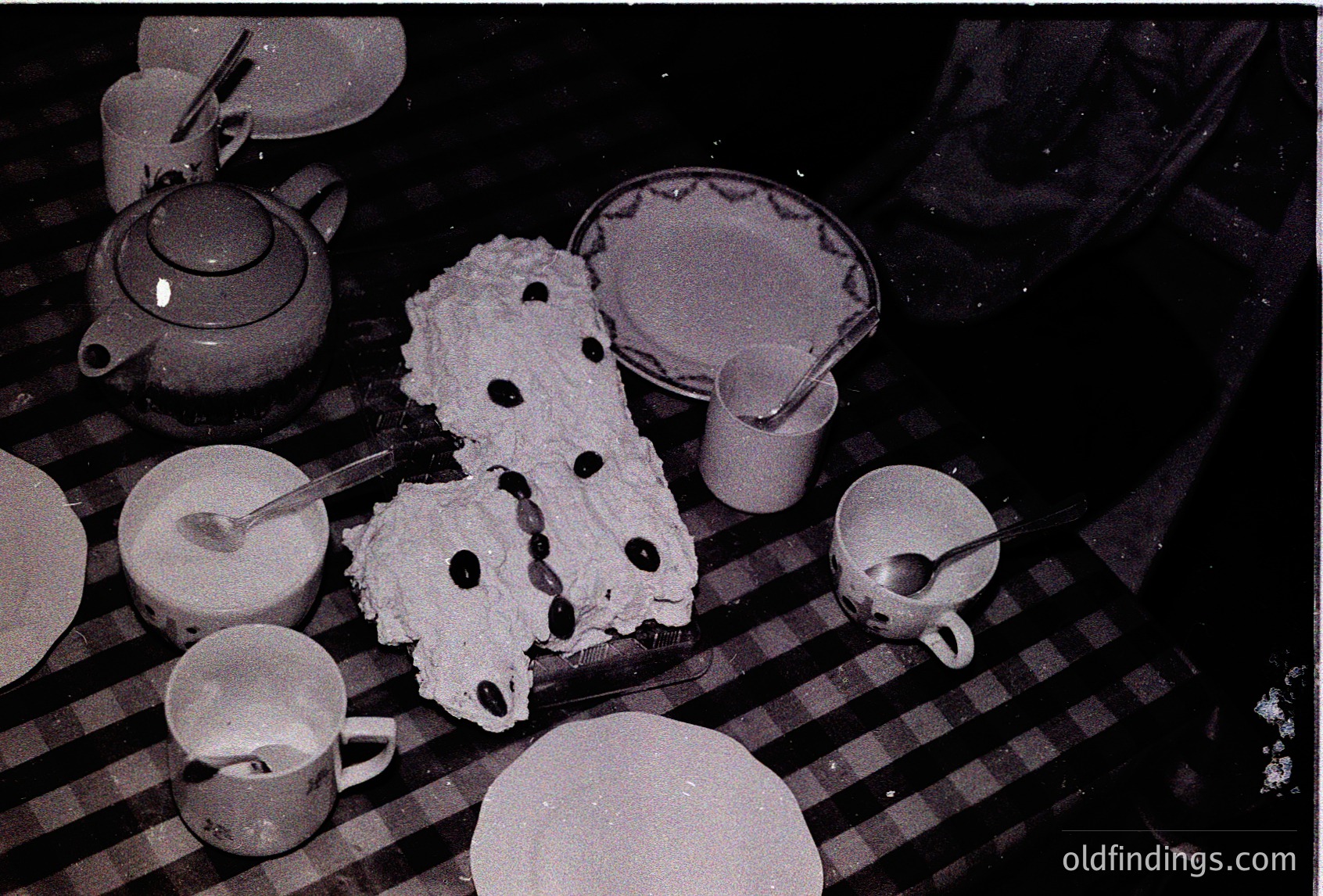 Mid-century dessert spread featuring layered sponge cake with jam filling, served on vintage crockery. White teapot, milk jug, and patterned plates suggest a 1950s–1960s European café setting. Warm, intimate dining ambiance captured in black-and-white. é