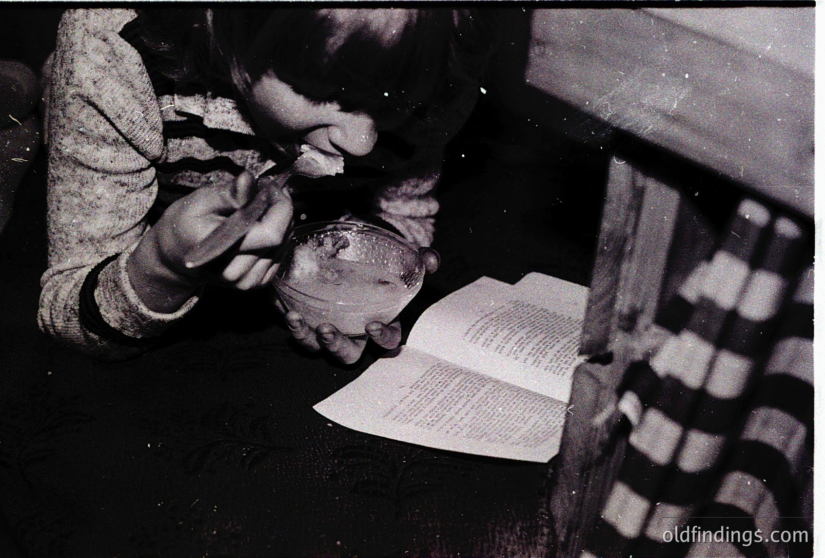 A child meticulously crafts with clay near an open book, likely a craft or art project. The scene suggests a mid-20th century domestic setting, possibly 1950s–1960s. The patterned wallpaper and vintage book hint at mid-century educational or family activities.