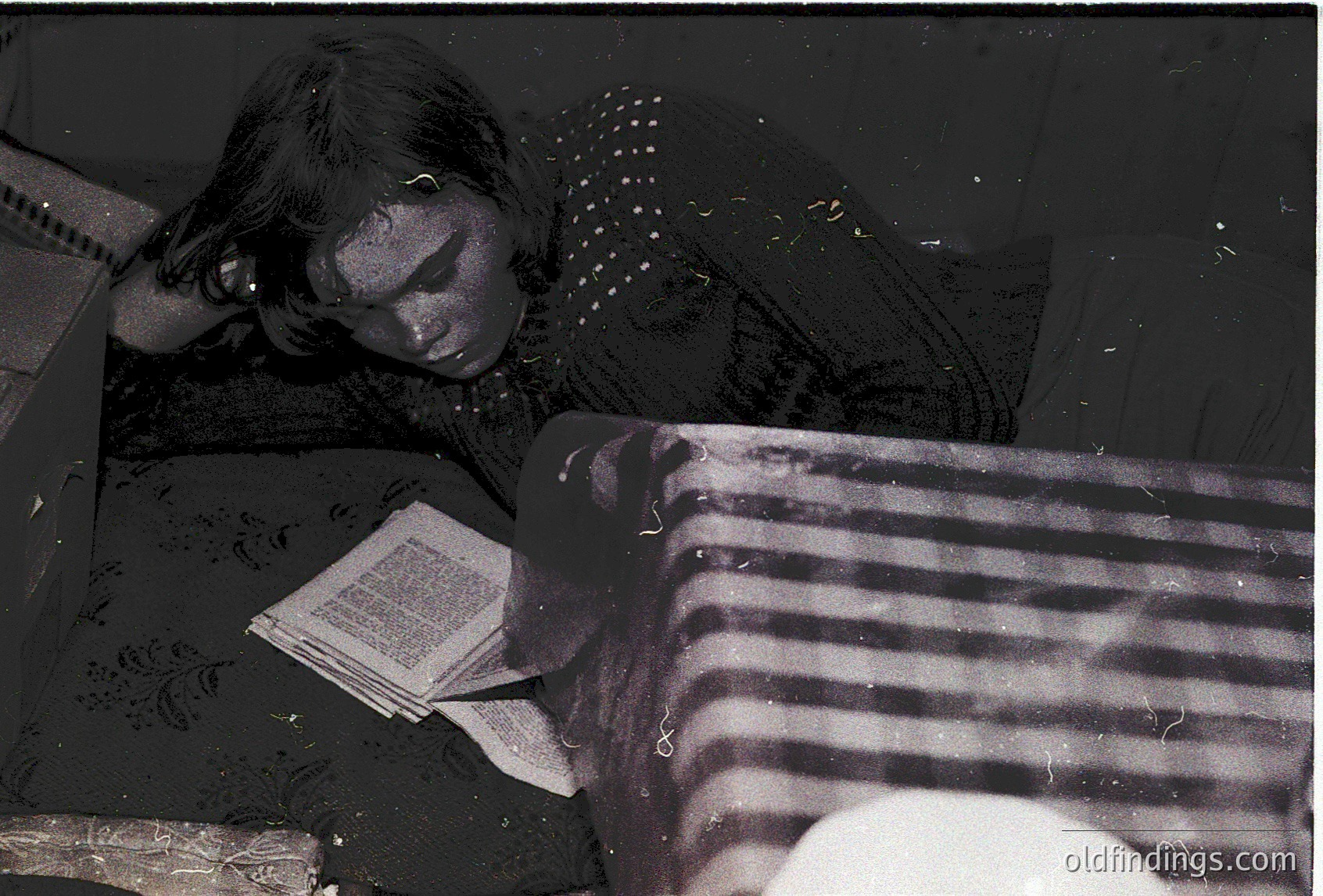 Vintage black-and-white photo of a child resting on a striped mattress, reading an open book. Patterned blanket and a round pillow visible. Likely mid-20th century domestic interior, evoking nostalgia for childhood reading habits.