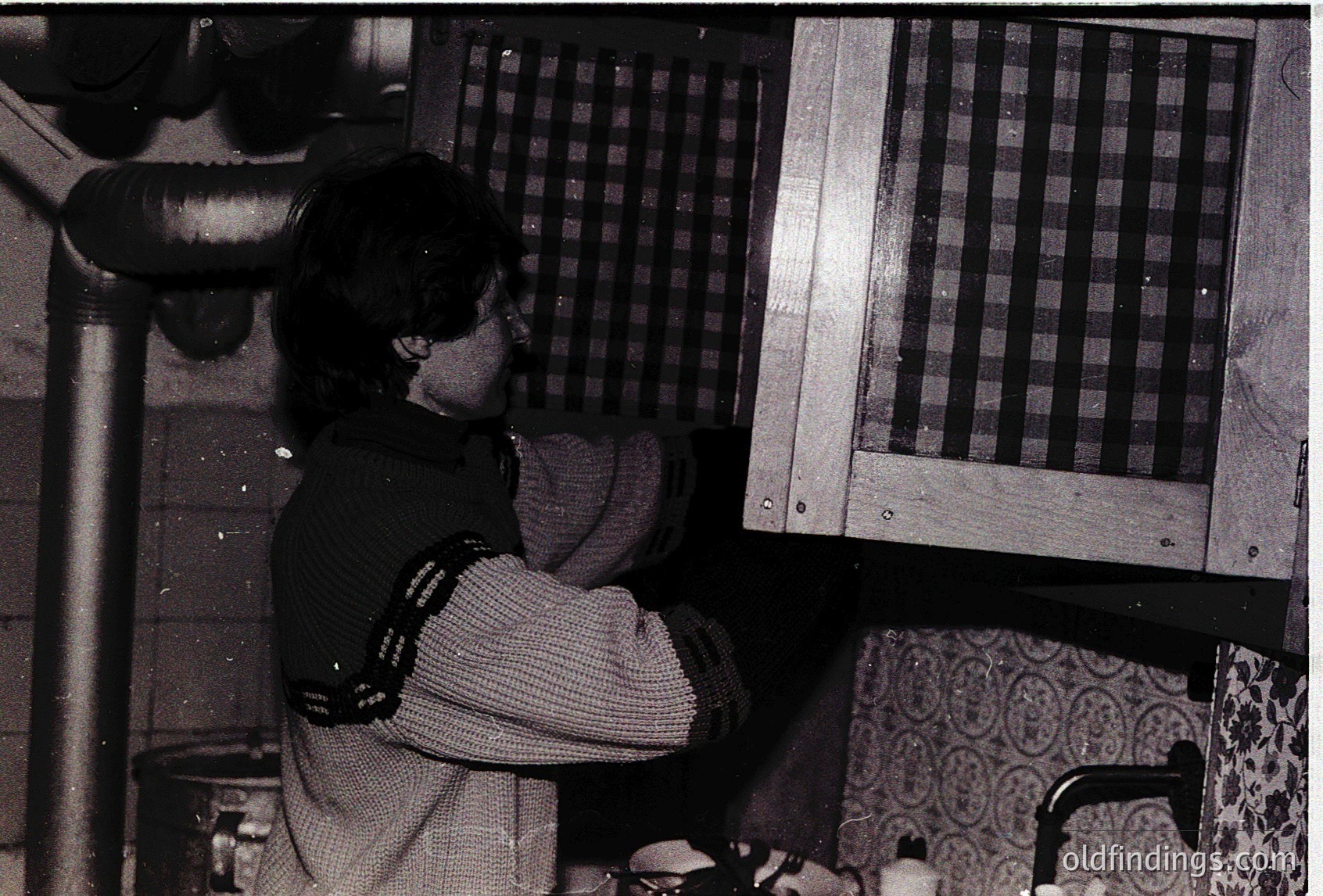 Vintage black-and-white photo of a person seated at a wooden table with a checkered-patterned wall behind them, likely a 1950s-1960s kitchen. The individual wears a striped sweater and holds a mug, with a pipe or stove visible on the left. Patterned fabric and a faucet suggest domestic life.