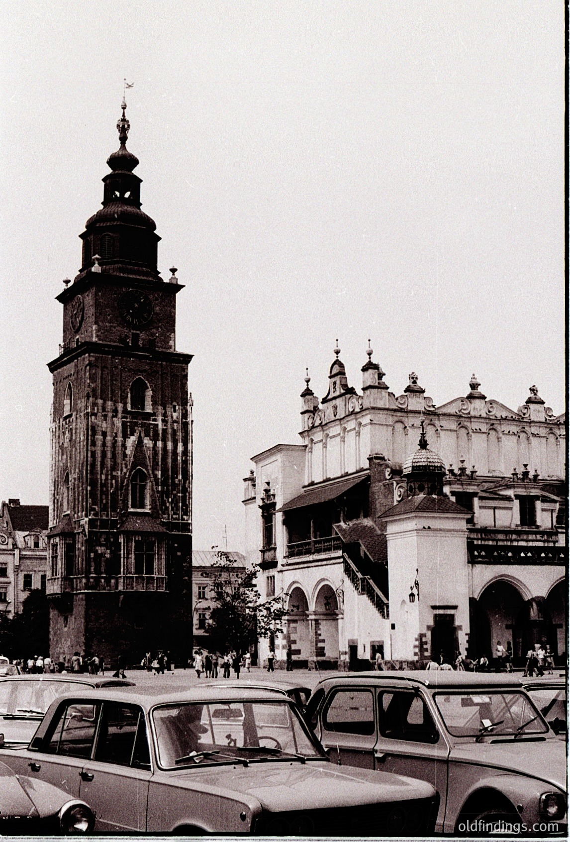 Historic 1960s black-and-white photo of Kraków’s **St. Mary’s Basilica** and its towering clock spire, flanked by Baroque-style church architecture. Mid-century cars and pedestrians populate the cobbled Main Market Square (). Distinctive Gothic-Renaissance hybrid design with ornate spire and arched windows.