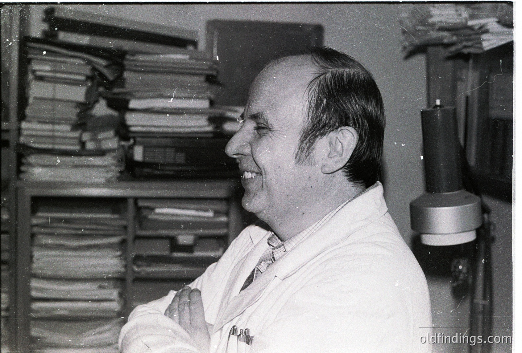 Mid-century academic in lab coat surrounded by stacked binders, likely a university or research setting. Classic 1960s–1970s office aesthetic with vintage typewriter in background.