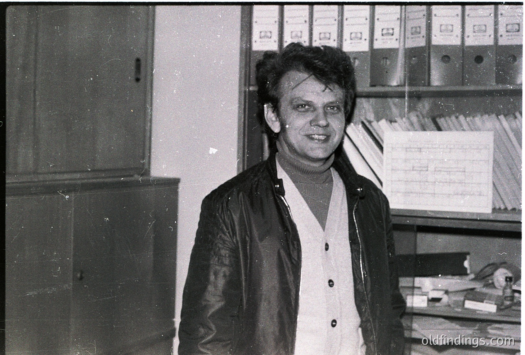Black-and-white office portrait of a man in a leather jacket and button-down shirt, standing beside metal filing cabinets and binders. Mid-20th century office environment suggests 1960s–1970s era.